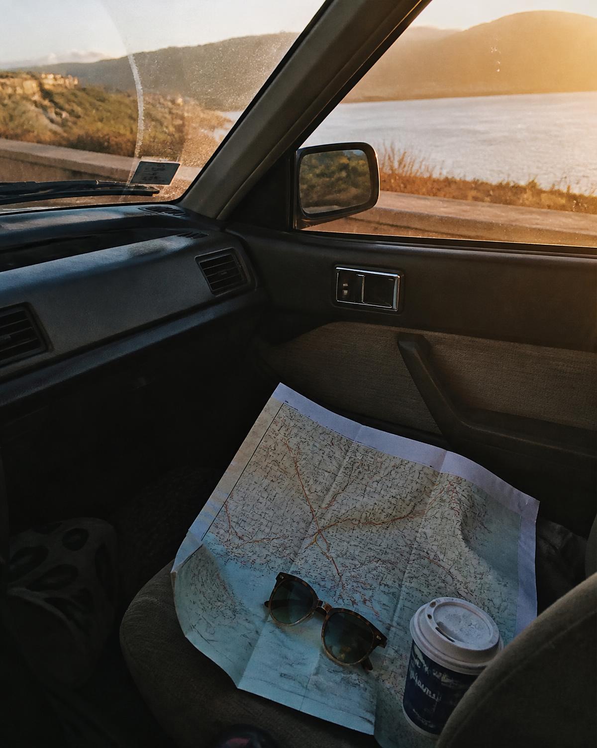 A folded paper road map, sunglasses and a coffee cup on a car seat at golden hour